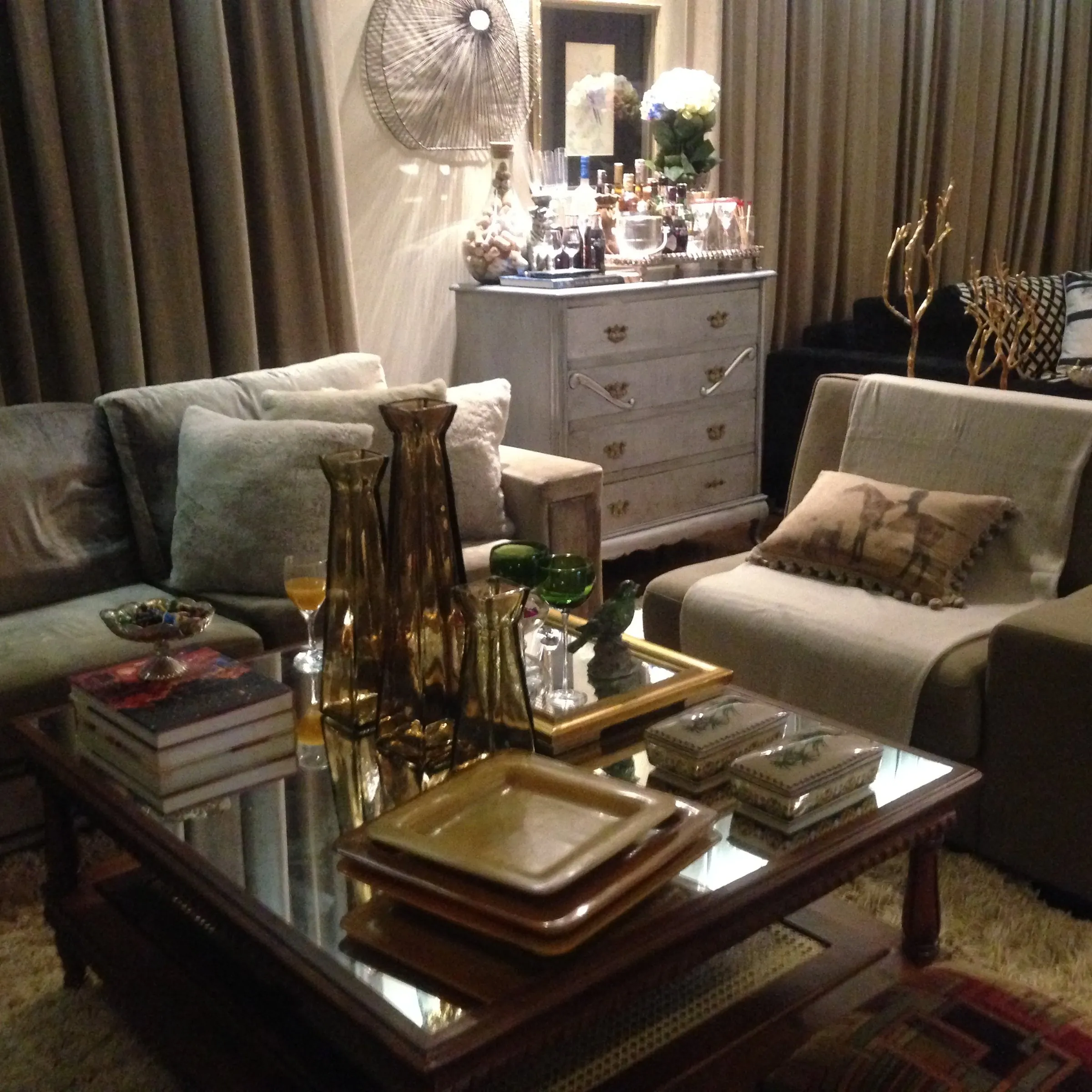 Sala com sofás claros, mesa de centro espelhada e cómoda branca com objetos